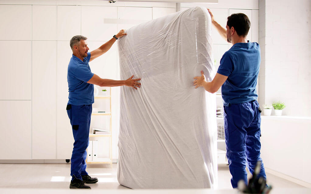 Two men wearing blue outfits maneuver a bed with a mattress cover on it inside of a home bedroom as they intend to move it out