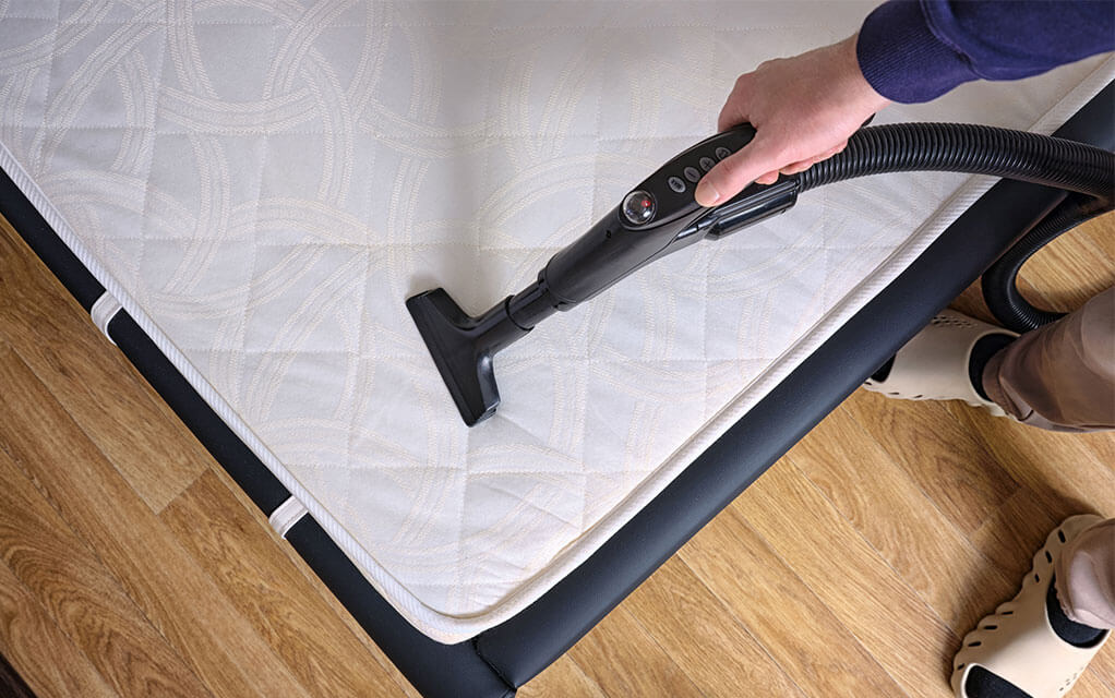 A person using a fabric cleaning vacuum cleans their pillow topper for their bed