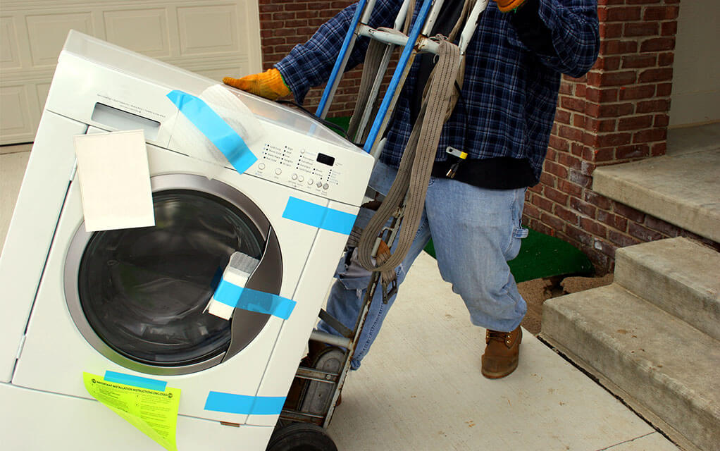 A professional laborer is moving a washing machine that is secure with tape and styrofoam as he wheels it out of a home with a moving dolly