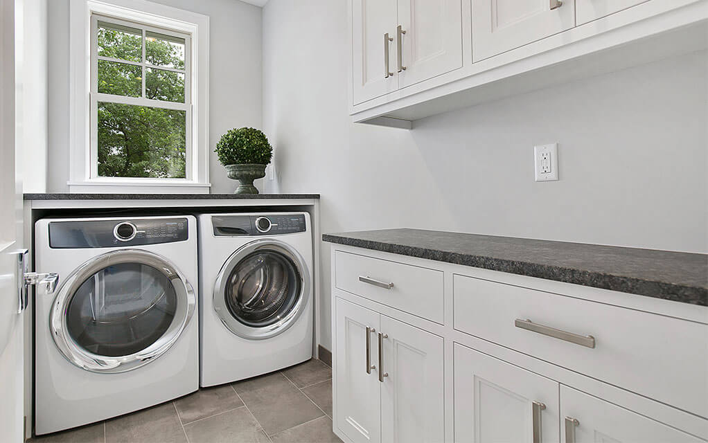 A view of an upscale home's laundry room, with a washer and dryer matching set sitting against the wall with a window over the top and an empty counter with cabinets above it
