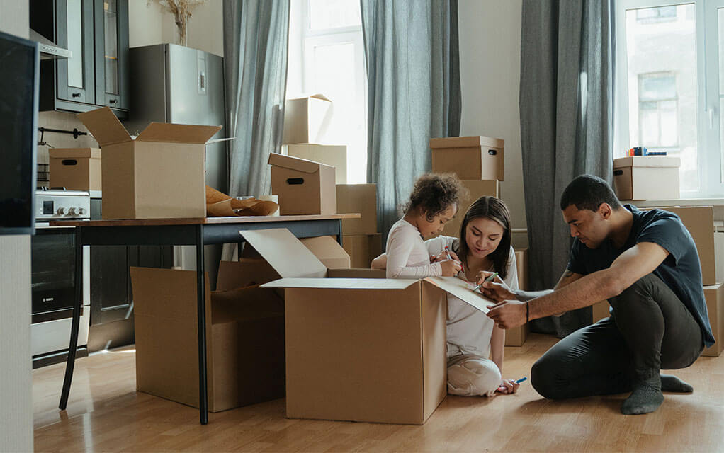 A young couple sit with their young daughter who is in a moving box as they play together, as boxes lay around them, ready to be moved and packed