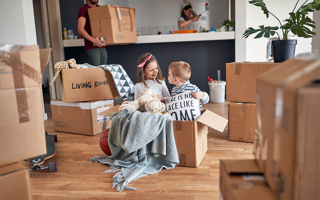 Two toddlers stand and play in a large moving box with some pillows and blankets inside in the living room as their parents are in the background packing and moving boxes around