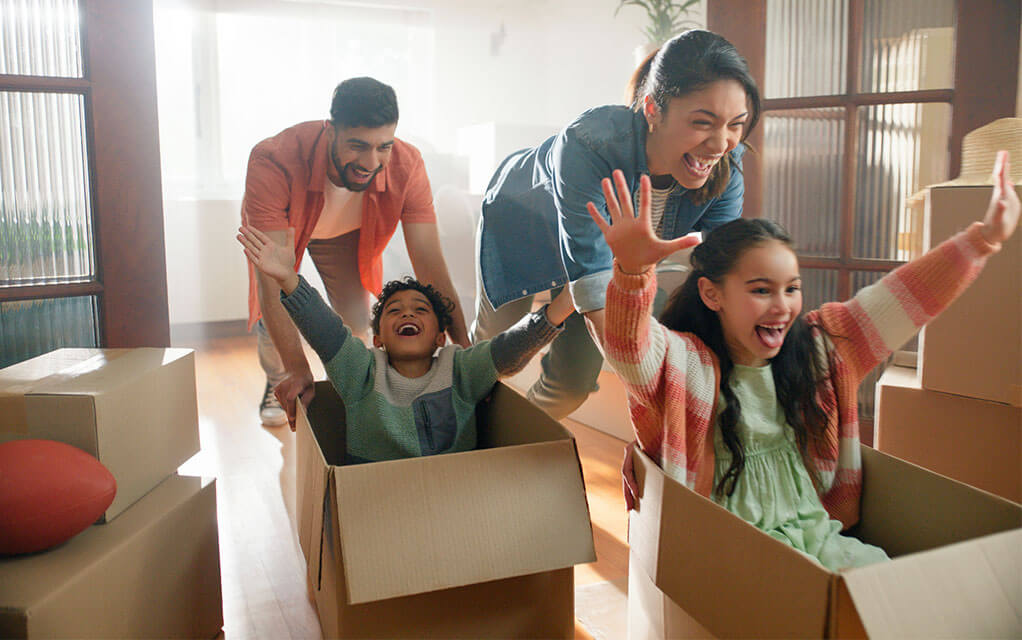 A mother and father push their two children around the home in open boxes, smiling, as their children laugh with delight and have their arms in the air