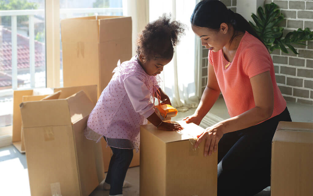 A mother helps her young daughter tape up a moving box, as other moving boxes lay open in the background in their living room on a sunny day