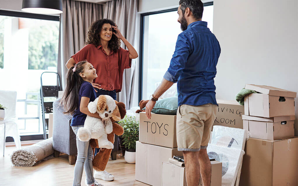 A middle aged couple is packing boxes and staging them in their home's living room as their young daughter holds a couple of stuffed animals
