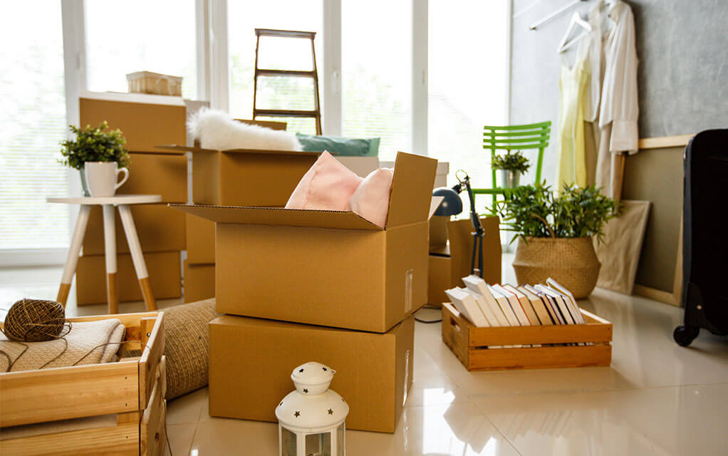 View of a number of boxes, packed and unpacked in a home's room ready to be packed away for an upcoming move, as books sit in an open crate on the floor with other house goods in view