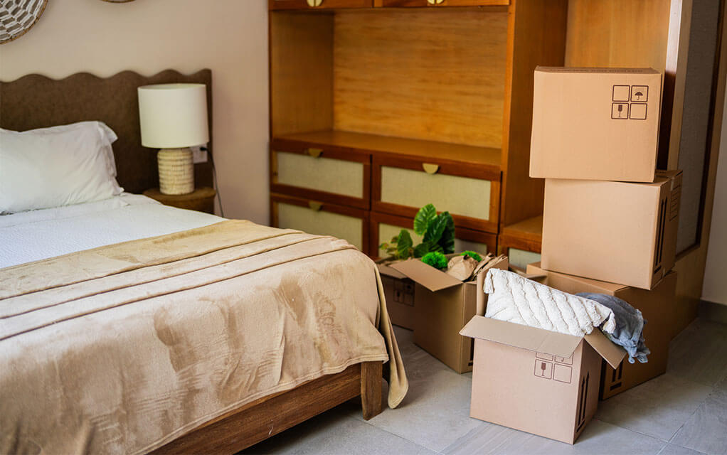 View of a bedroom, with a made bed and moving boxes, both packed and unpacked and open waiting to be closed up for an upcoming move