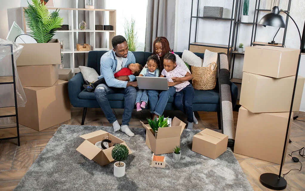 A young family all sit on a couch together looking at something on their laptop as moving boxes - packed and unpacked - sit around them in their home