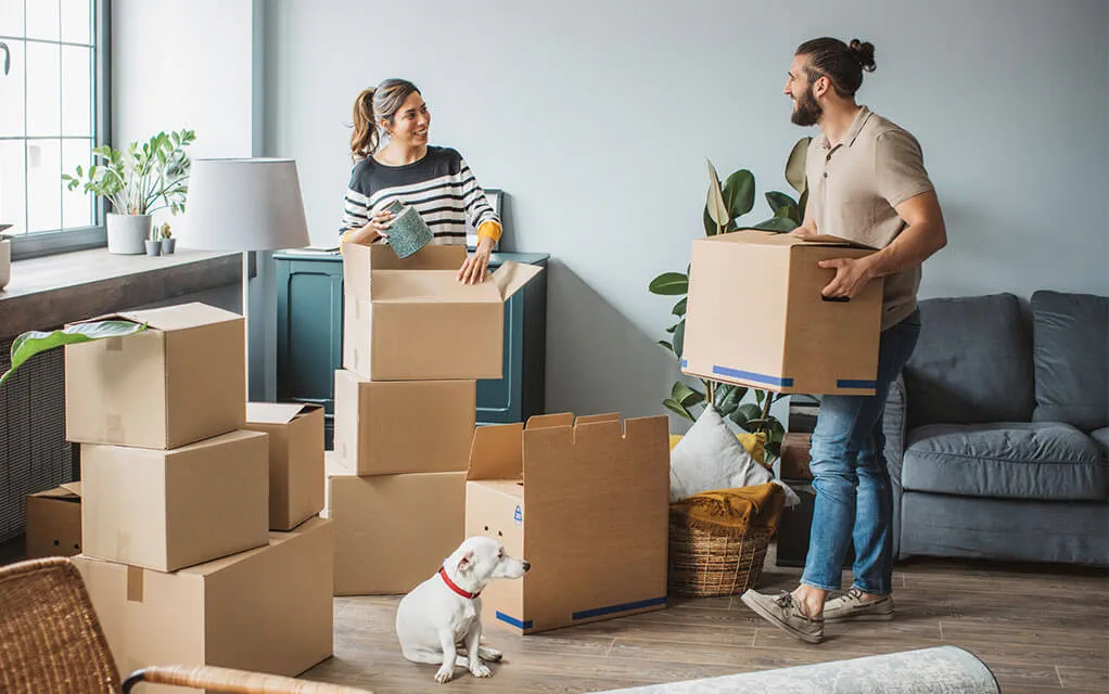 A young couple carries boxes into their new home, as boxes and furniture sit on the floor around them as their small dog looks up at them