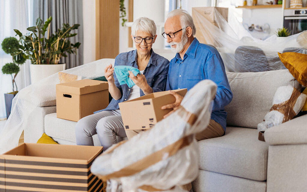 A senior couple sit on their couch in their living room, taking things out of moving boxes as their belongings sit around them, waiting to be unpacked
