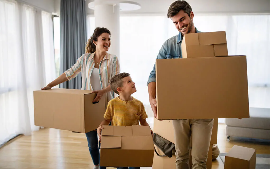 A happy couple and their young son are all smiling and laughing as they carry moving boxes out of their home on a sunny day