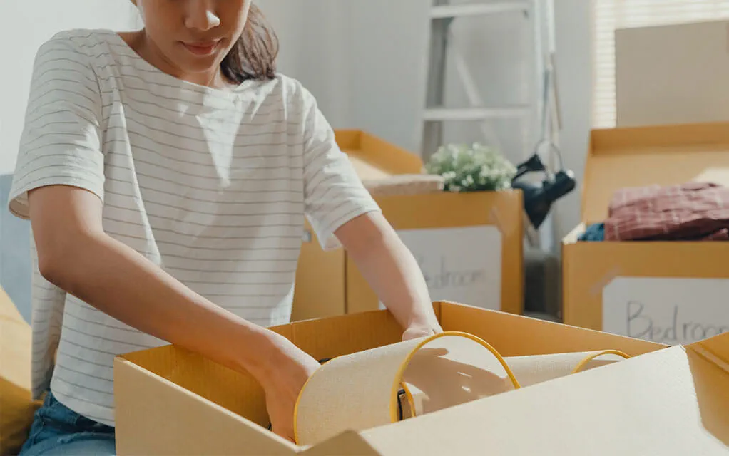 A woman is placing a lamp and its shade into a box for an upcoming move, as other boxes sit open in the background with the word "Bedroom" written on them