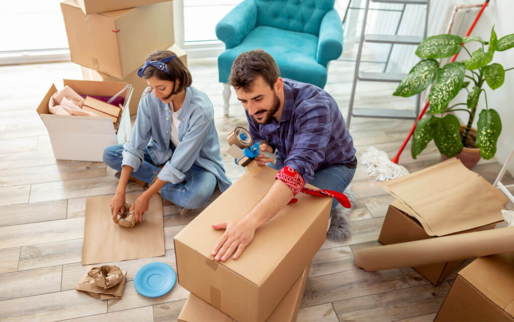 A happy couple sits on the floor of their house, packing away their belongings and fragile items into tissue paper and boxes as they get ready for their upcoming move