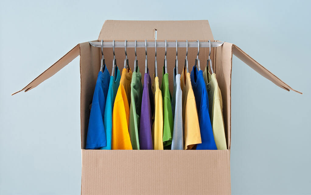 View of an open wardrobe moving box, with colorful shirts all sitting on the box's hanging rod inside