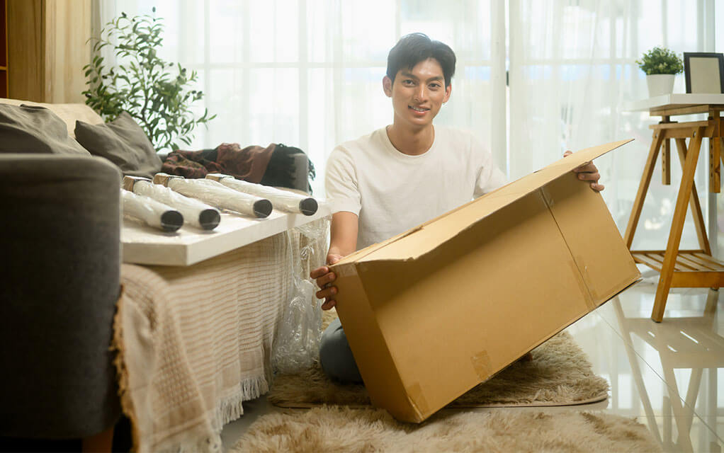 A young man holds open a box as he sits on the floor of his living room, ready to pack away some artwork inside for safekeeping