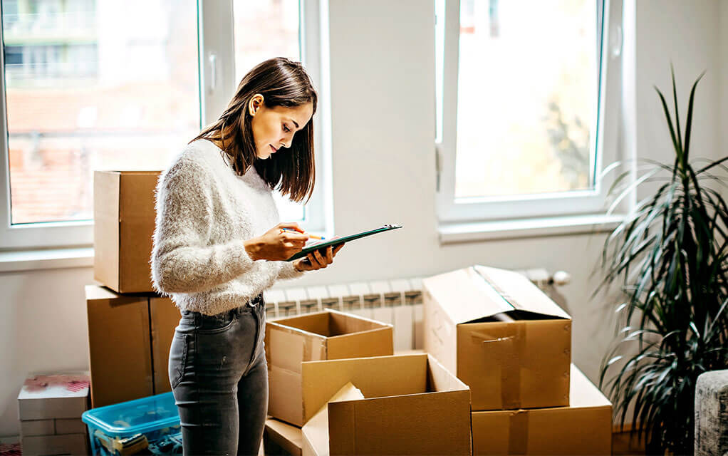 A woman jots down notes on a pad of paper, as opened moving boxes sit on the floor in the background