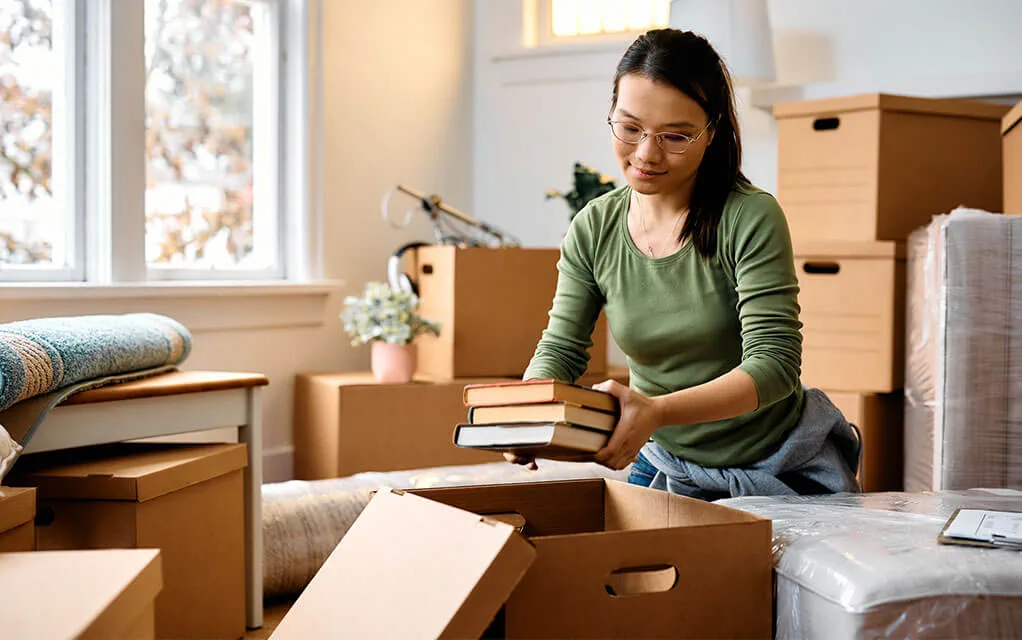 A woman in a green shirt packs books away into a moving box as other closed boxes lay on the floor around her