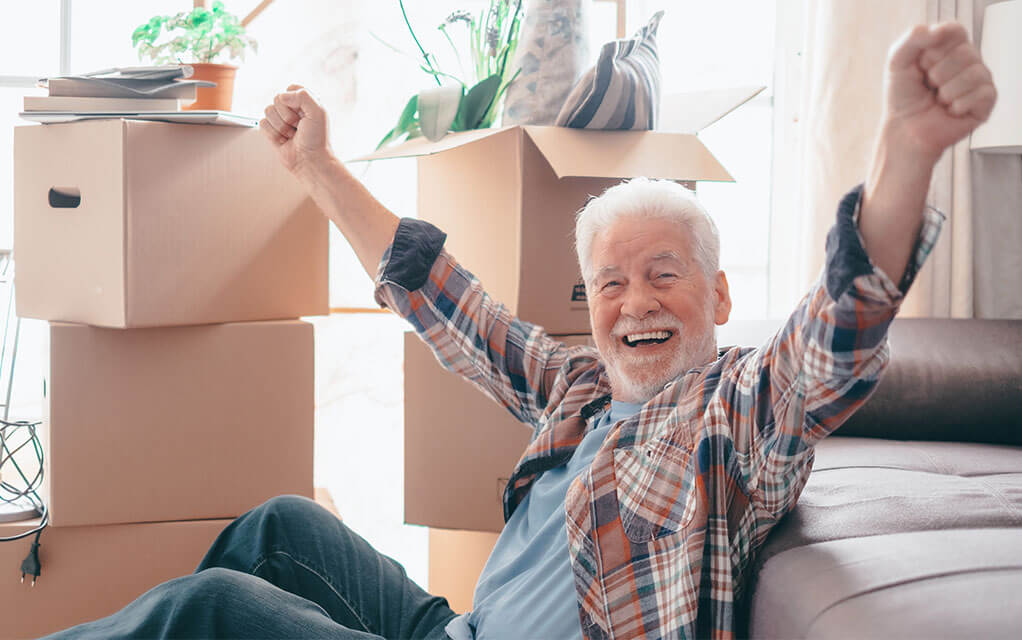 An older man smiles and holds his arms up in celebration while sitting on the floor in front of his couch with packed moving boxes staged in the background