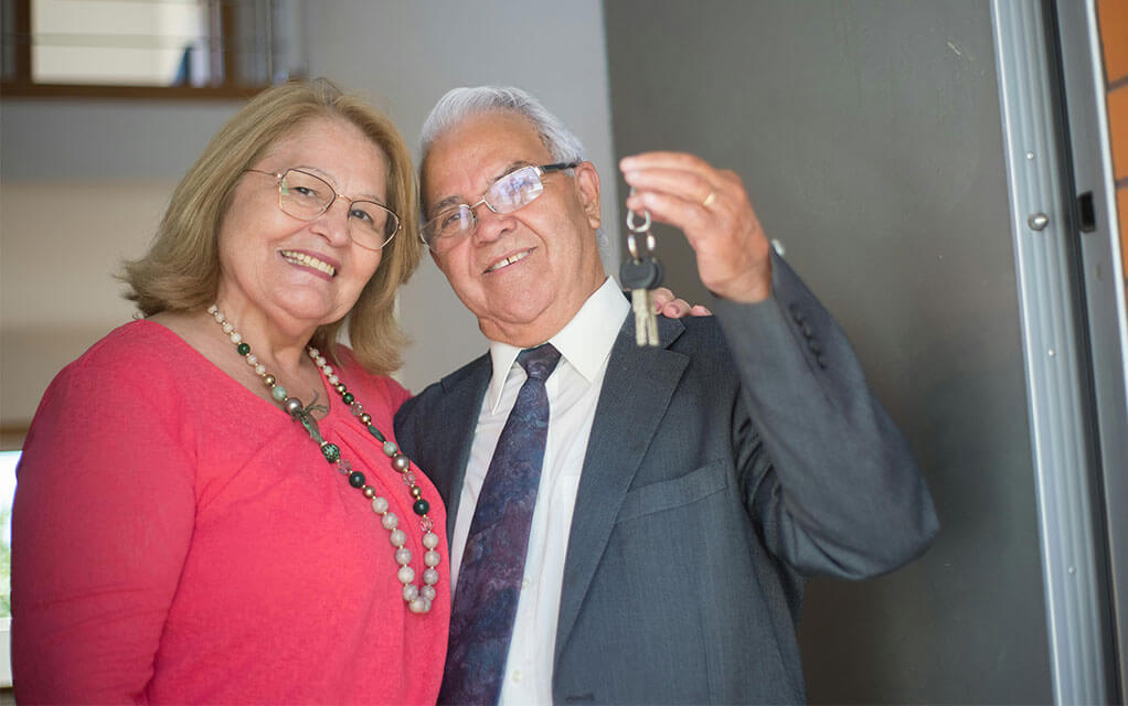 A senior couple poses for a picture in the doorway of a home while the man holds a pair of house keys in his hand