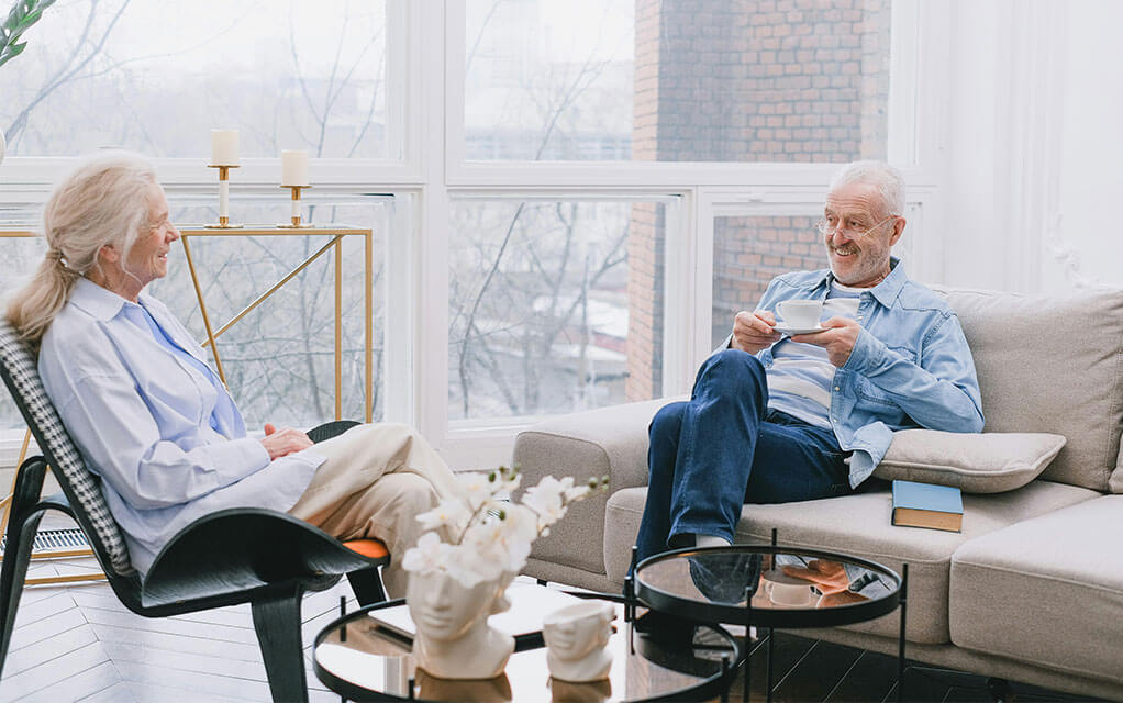 A senior couple sit in their home's living room having a pleasant conversation while the man drinks coffee on a cloudy day