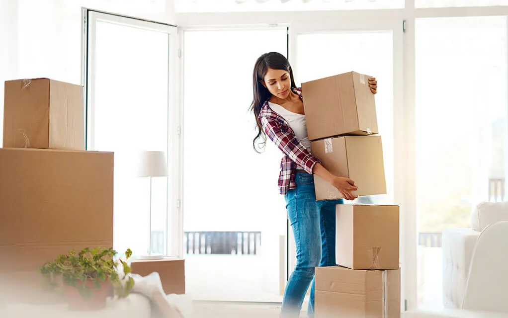 A young woman holding two small moving boxes stacks them on top of two other moving boxes on the floor, as her patio door lays open behind her on a sunny day