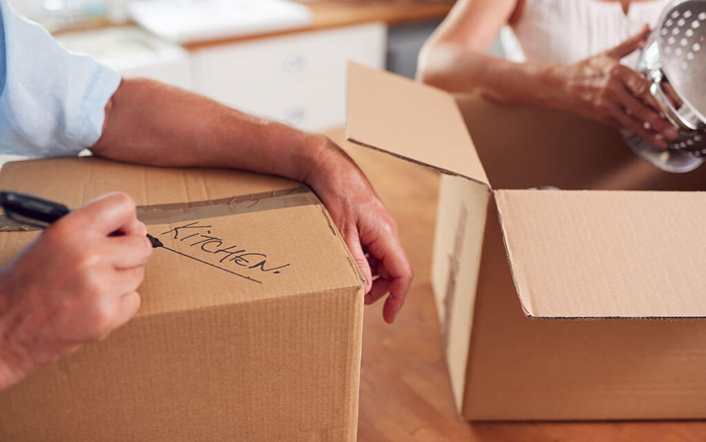 Two people are packing moving boxes in their kitchen, as one person writes "kitchen" on one of the boxes with a marker
