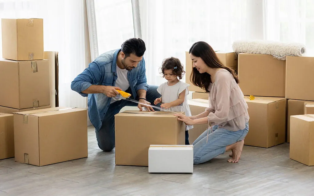 A young couple and their small daughter close up a moving box with some tape, surrounded by more moving boxes waiting to go to their new home