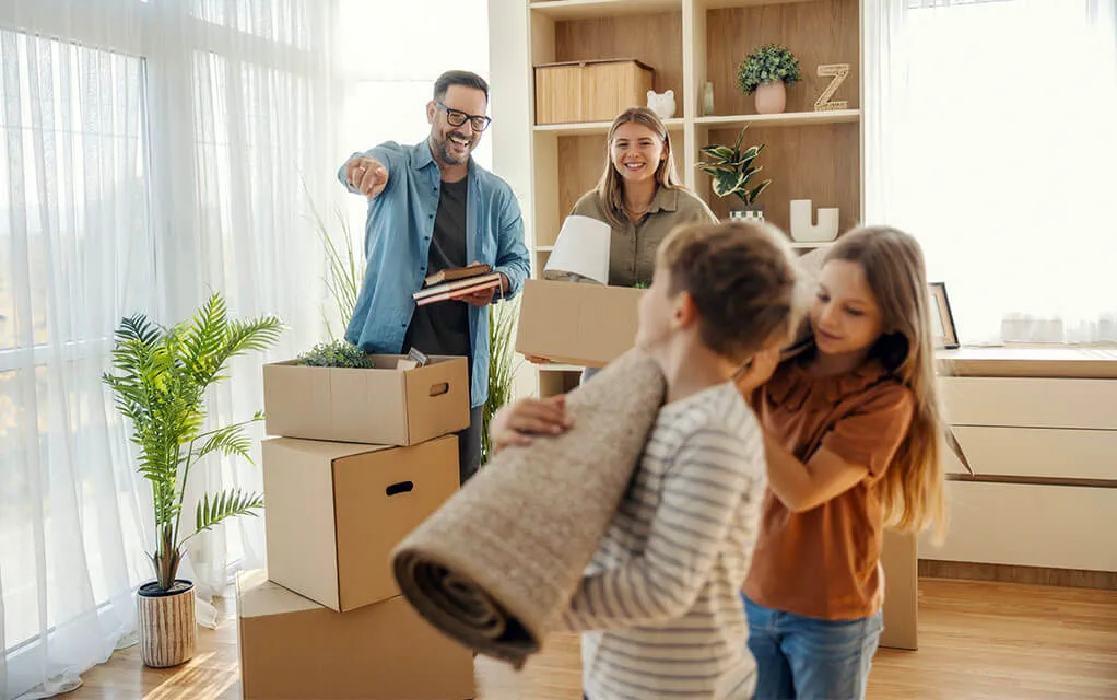 A mother and father laugh and smile as their children help carry a rolled up rug into another room as they move into their new home