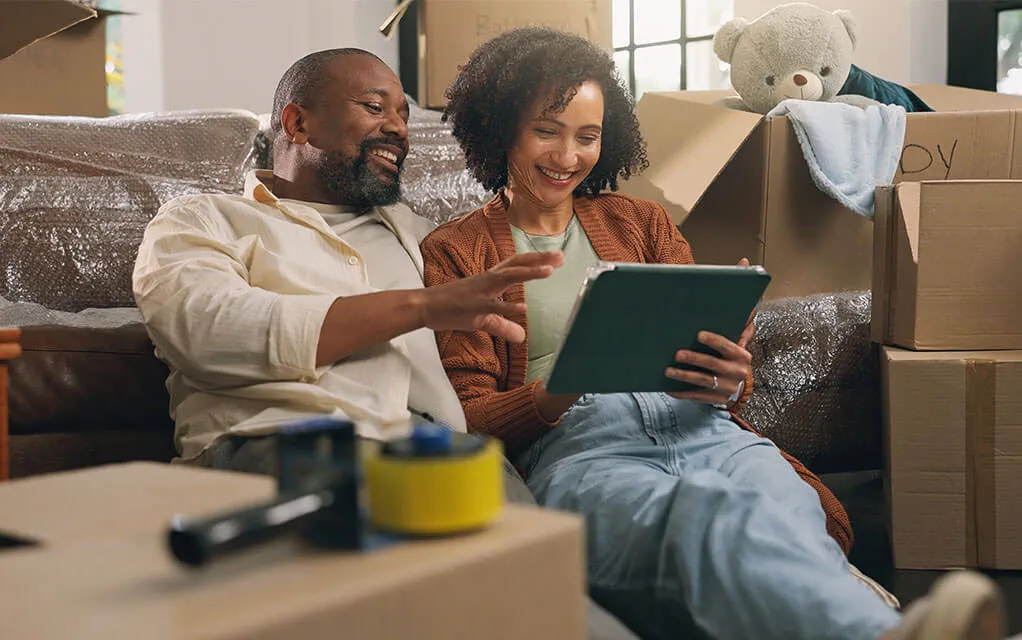 A middle aged couple sits on the floor in front of their covered couch, surrounded by moving boxes as they look at something excited on their tablet