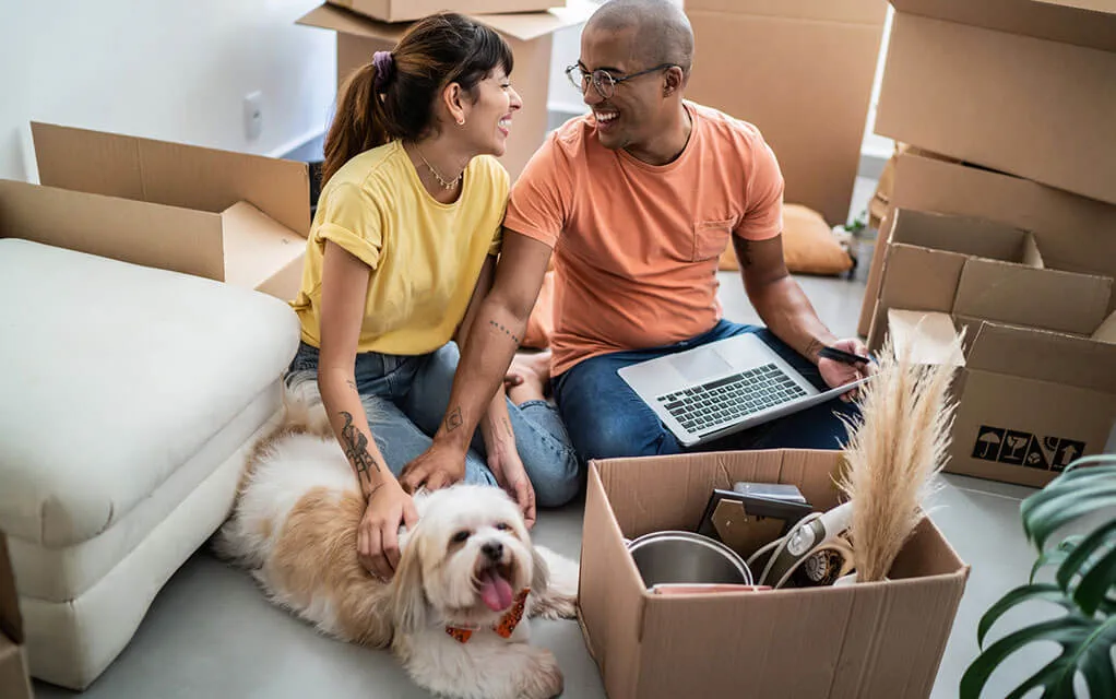 A couple sits on the floor with their dog with a laptop and an open moving box with their stuff in it while more moving boxes sit in the background