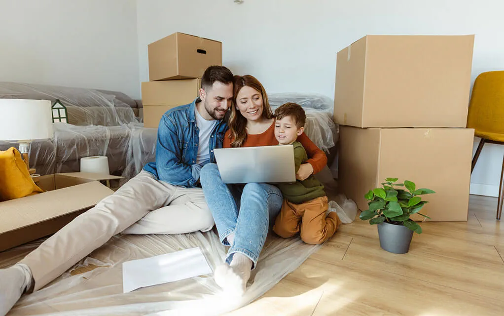 A happy young couple and their child all sit on the floor of their new home surrounded by boxes while looking at something on their laptop computer