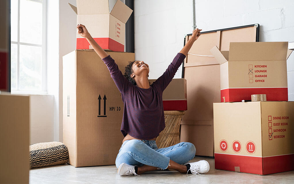 A young woman has her arms up in the air in a victory pose as she is surrounded by moving boxes, both opened and closed