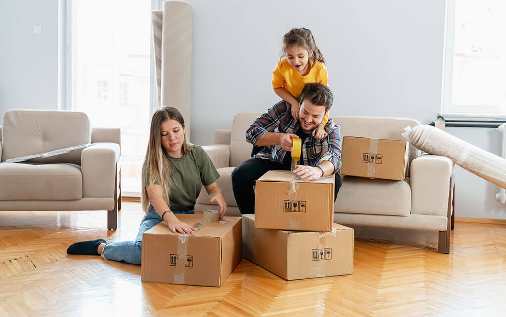 A young family closes moving boxes with tape in their old home's living room as they prepare for an upcoming move
