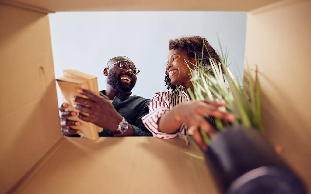 A view from the bottom of a moving box shows a couple placing items such as a small plant and books inside of it