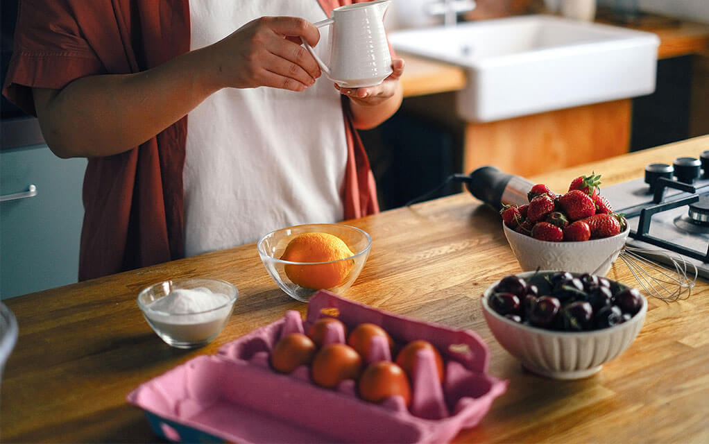 A person stands at their kitchen island as berries, eggs, an orange, and sugar in a bowl sit on the island in front of them as they hold a cup in their hands