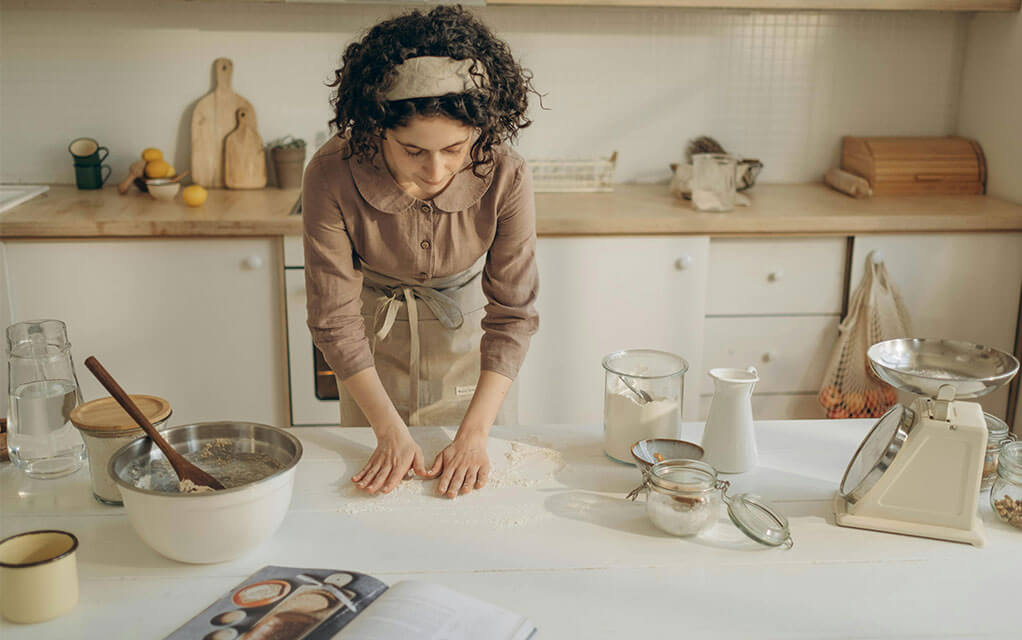 A woman is beginning to create dough on her kitchen island as mixing bowls, ingredients in jars, and a cook book lay open in front of her
