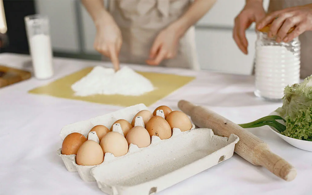 A dozen organic eggs sit in a carton on a white marble kitchen island waiting to be used as two people begin to create pasta in the background