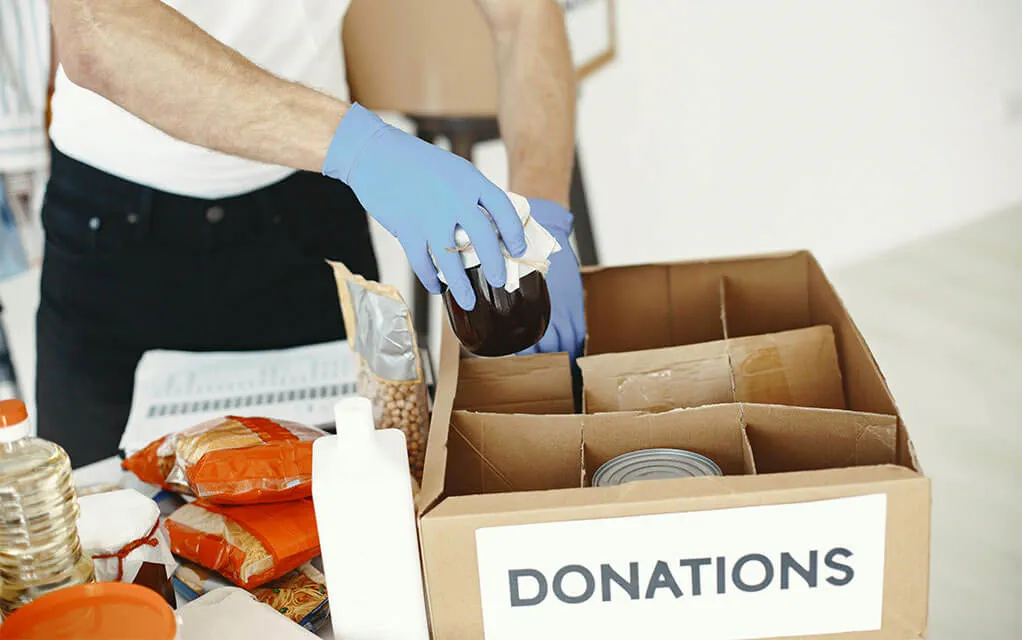 A person wearing blue gloves places jars and cans into a box with dividers in it labeled "DONATIONS" as other foodstuffs sit next to it waiting to be sorted