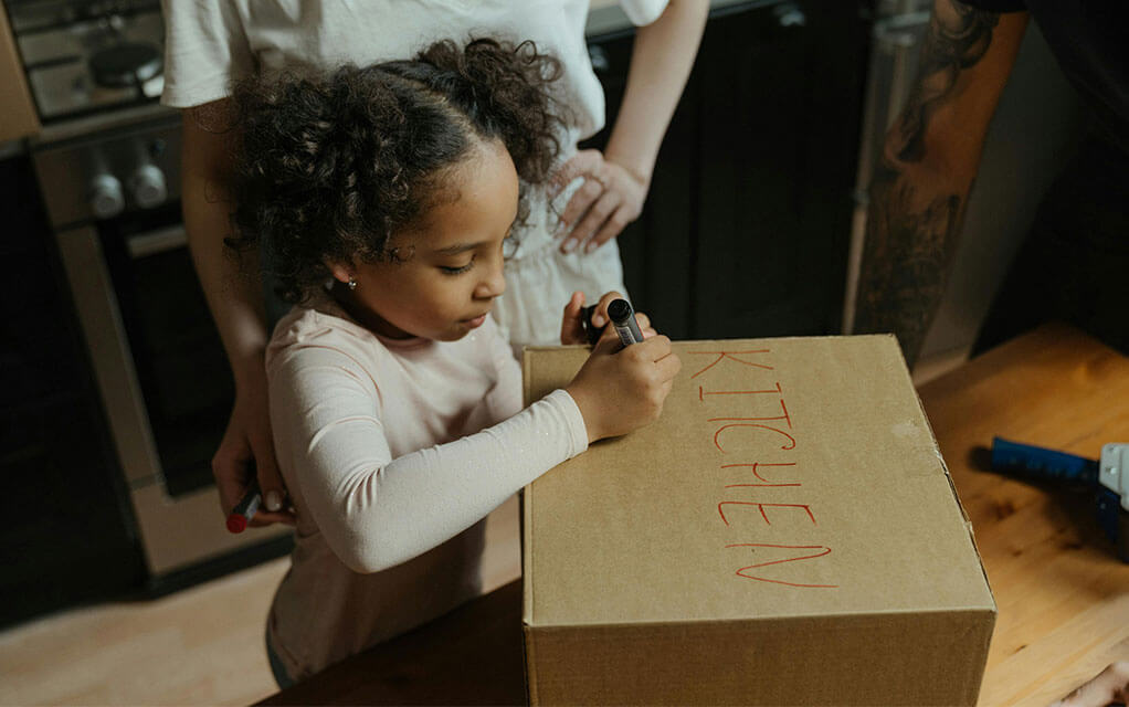 A small child marks a moving box with the word "KITCHEN" written on it as her parents watch her