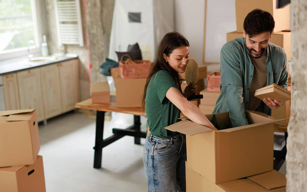 A happy and smiling couple packed books and other belongings into an open moving box sitting on top of other boxes as they pack up their home for a move on a sunny day