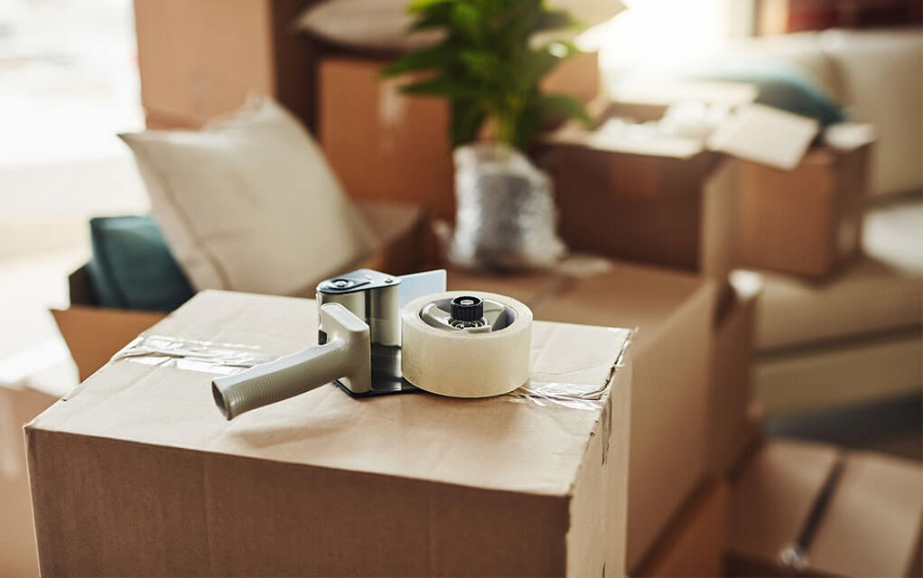 A tape gun with some tape on it lays on a taped up and ready to be packed box, waiting to be used again as other packed belongings and boxes sit in the background