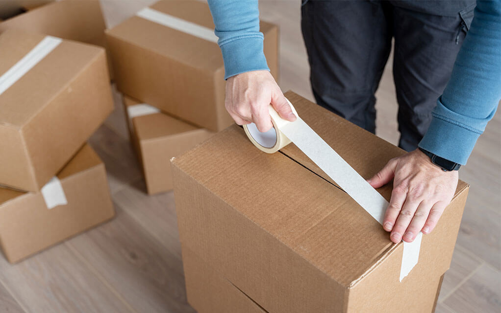 A man uses a roll of tape to secure the top of a moving box he has just finished packing. In the background behind him, other packed and taped up boxes sit waiting to be moved