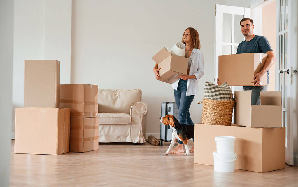A young couple carries boxes into their new home as their dog walks in with them - boxes already sit on the floor that have been brought inside