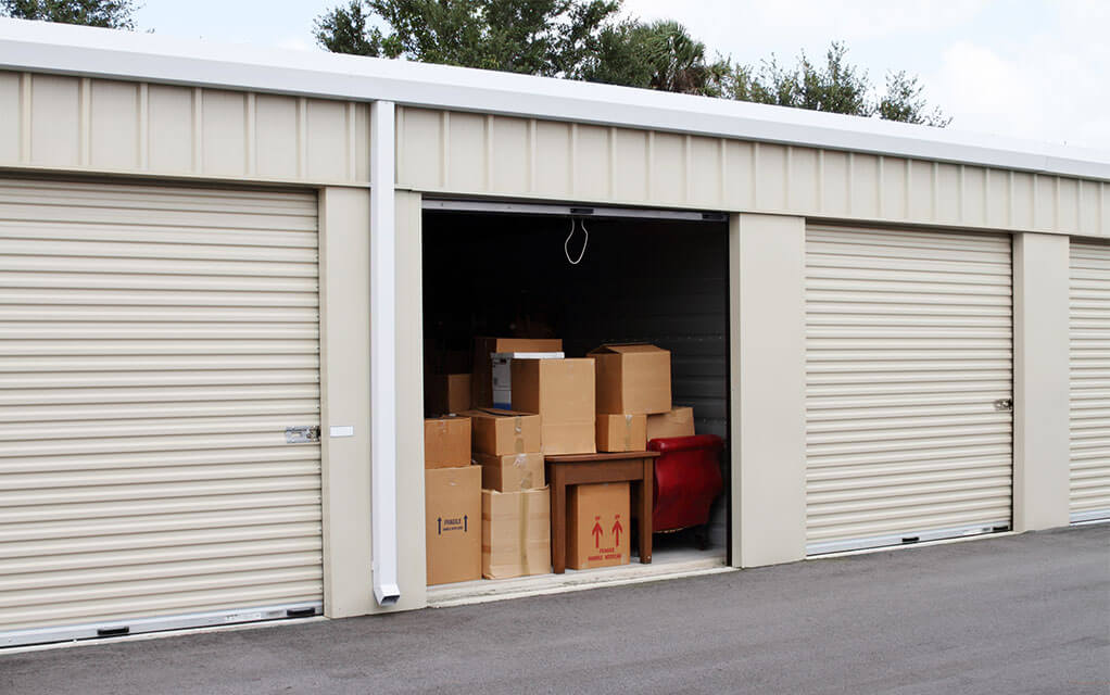 View of an open storage unit filled with boxes and furniture in between two closed units at a traditional self-storage facility on a cloudy day