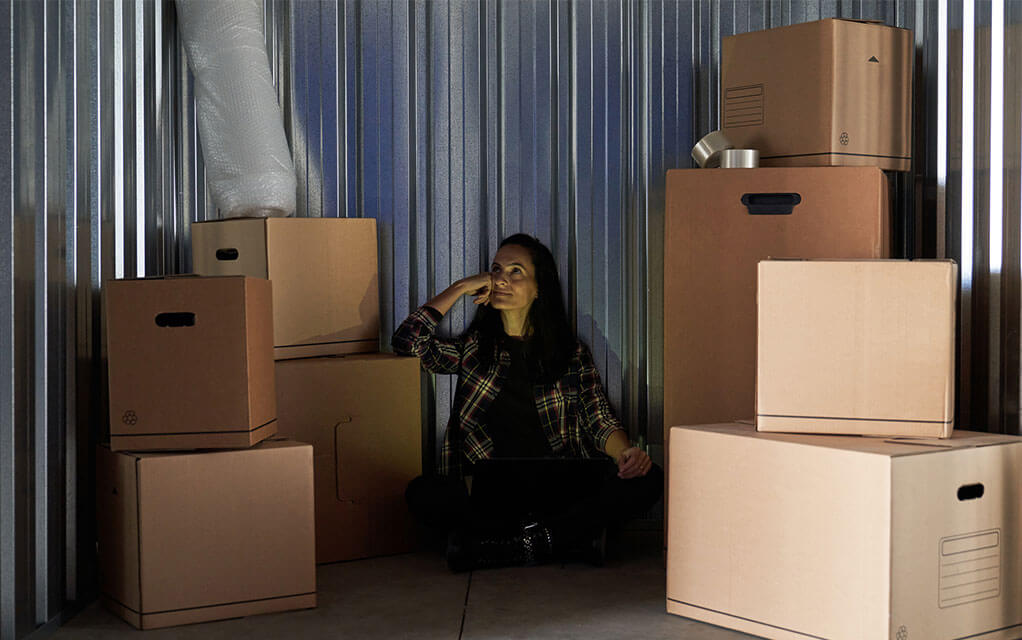 A slightly annoyed woman sits in the middle of a dark storage unit, with a laptop sitting in her lap as boxes lay stacked around her in the storage locker