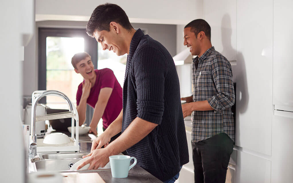 Three young male roommates laugh as one of them washes dishes in the sink of their apartment on a bright day