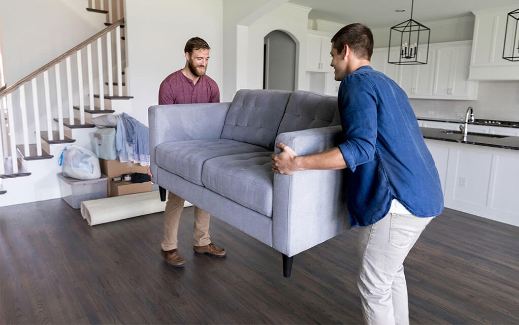 Two men are moving and positioning a loveseat in a home's living room as other unpacked and unplaced items lay on the floor in the background next to the stairwell