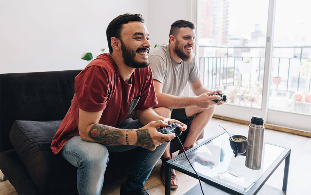 Two male roommates sit on their apartment couch in their living room, playing video games and laughing together on a bright afternoon day