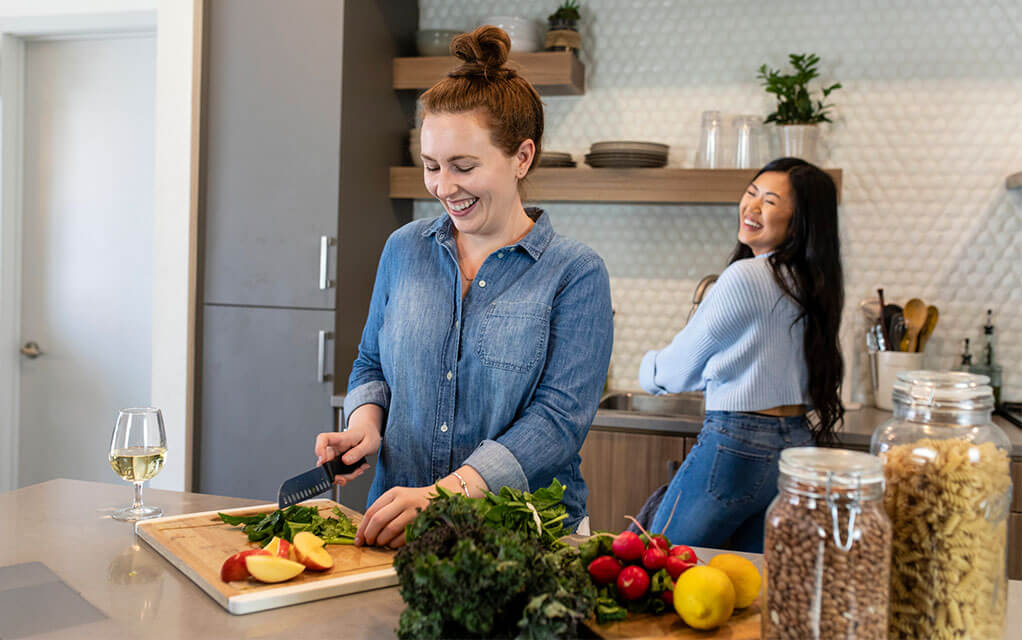 Two young women and roommates laugh as they make and prepare food in the kitchen of their apartment