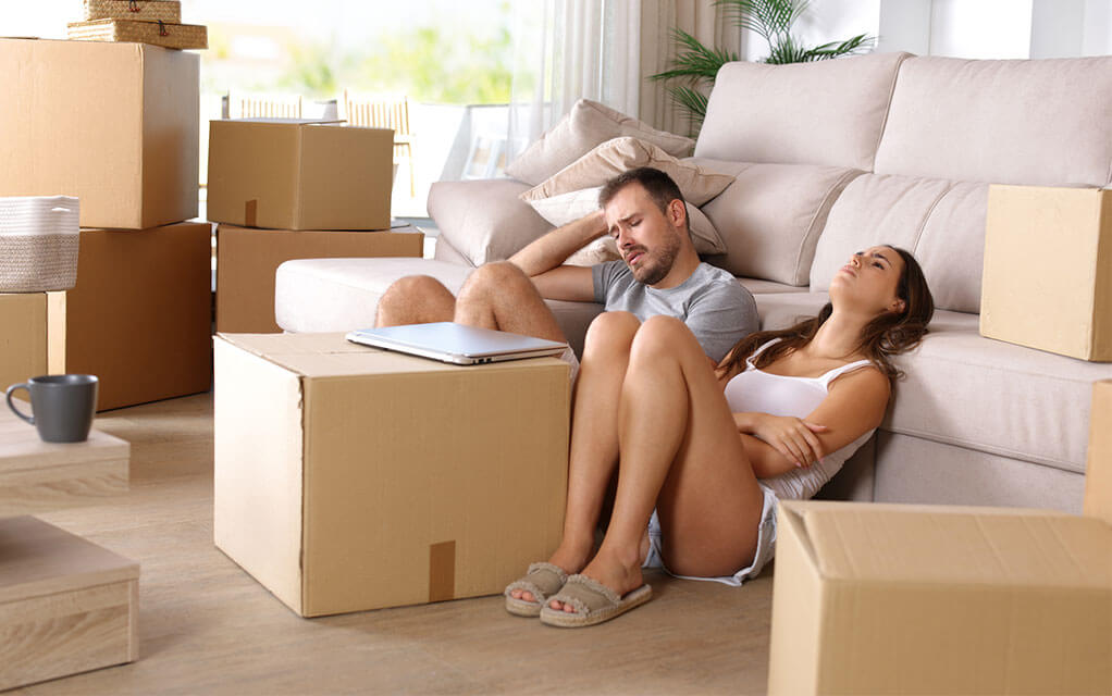 A frustrated and upset young couple sits on the floor of their home against their couch, surrounded by packed moving boxes as their laptop sits closed on a moving box in front of them
